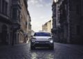 A white Porsche Macan Electric with headlights on is parked on a cobblestone street between tall, historic buildings at dusk—capturing the spirit of a Road to Christmas journey.