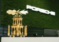A wooden Christmas pyramid with lit candles sits on a counter in front of a green wall featuring the illuminated Porsche logo, highlighting the Road to Christmas spirit alongside the innovation of Porsche Macan Electric.