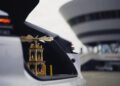 A wooden Christmas pyramid sits in the open trunk of a Porsche Macan Electric, with a modern, circular building in the background—capturing the spirit of the Road to Christmas.
