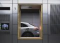 A silver Porsche Macan Electric is charging at an indoor station, viewed through a square window in a tiled wall—capturing a quiet moment on the Road to Christmas.