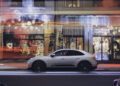 A white Porsche Macan Electric SUV is parked on a city street at night in front of shops decked with Christmas lights and ornaments, with blurred light trails from passing traffic, capturing the spirit of a Porsche Christmas.