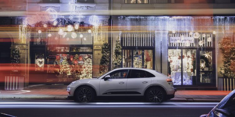 A white Porsche Macan Electric SUV is parked on a city street at night in front of shops decked with Christmas lights and ornaments, with blurred light trails from passing traffic, capturing the spirit of a Porsche Christmas.