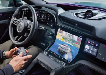 A person sits in a Porsche Cayenne’s driver seat holding a game controller, with a racing video game displayed on the car’s central touchscreen, blending immersive driver experience with digital entertainment.
