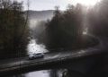 A white electric car drives across a bridge over a river surrounded by trees on a misty morning with sunlight in the background, evoking a serene scene reminiscent of The Road To Christ.