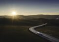 A Porsche Macan Electric drives on a winding road through open countryside at sunset, with the sun low on the horizon and rolling hills in the background.