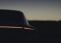 A Porsche Macan Electric's rear end with a horizontal LED taillight illuminated, photographed at dusk against a dark, blurred background.