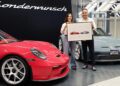 A man and woman stand between a red and a grey Porsche 911 S/T, holding a framed sketch of the two cars, in front of a sign that reads "Sonderwunsch.