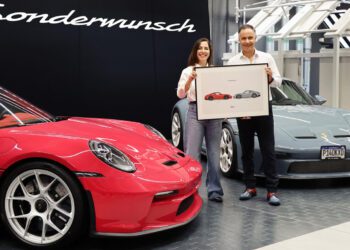 A man and woman stand between a red and a grey Porsche 911 S/T, holding a framed sketch of the two cars, in front of a sign that reads "Sonderwunsch.