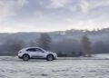 A white Porsche Macan Electric drives along a frost-covered country road with a blurred background of trees and hills under a cloudy sky.