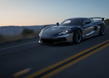 A sleek black Rimac Nevera R hypercar drives at high speed on an empty highway at sunset, with blurred surroundings indicating motion.