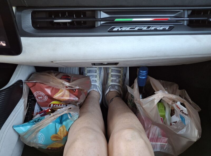 Person sitting in a car passenger seat with their legs extended, surrounded by grocery bags filled with snacks and drinks—proving that even on a luxury getaway, can a 200-mph supercar really pull off style and comfort?.