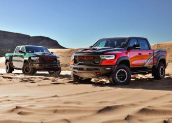 Two Ram TRX pickup trucks, one green and the other black and red, are parked on sandy dunes. These gas pickups boast up to 777 HP, standing out against scenic hills and a clear blue sky in the background.