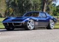 A dark blue Corvette Restomod classic Chevrolet sports car is parked on a paved road with trees and grass in the background, as seen at Mecum Kissimmee supporting Curing Kids Cancer.