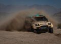 A beige D7X-R off-road vehicle speeds through a dusty desert trail, kicking up a large cloud of sand, with mountains visible in the background.