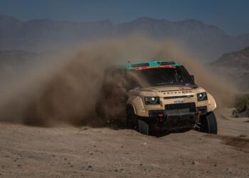 A beige D7X-R off-road vehicle speeds through a dusty desert trail, kicking up a large cloud of sand, with mountains visible in the background.