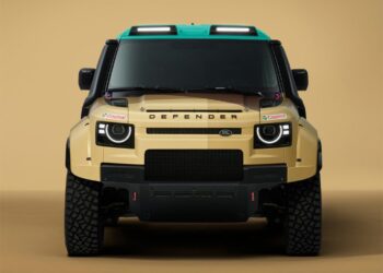 Front view of a tan Land Rover Defender with rugged tires, black accents, a green roof, and Castrol sponsor stickers, set against a beige background.