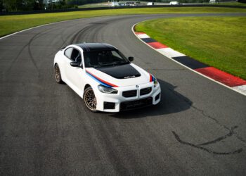 A white BMW coupe with racing stripes is parked on a paved race track near a grassy bend.