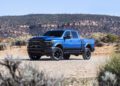 A blue 2027 Ram Power Wagon pickup truck is parked on a dirt road in a desert landscape with shrubs and rocky hills in the background, ready to showcase its impressive torque.