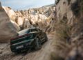 A Porsche Macan Electric navigates a narrow, rocky dirt road in Cappadocia, Turkey, surrounded by large beige rock formations and sparse vegetation.